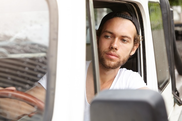 Gratis foto sluit omhoog geschoten van modieuze jonge ongeschoren mens die glb achteruit draagt, binnen zijn jeep zit en weg bekijkt terwijl het parkeren van zijn auto met vierwielaandrijving