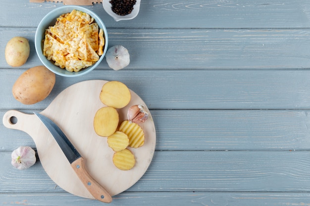 Sluit omhoog mening van aardappelplakken en knoflook met mes op scherpe raad en chips op houten achtergrond met exemplaarruimte