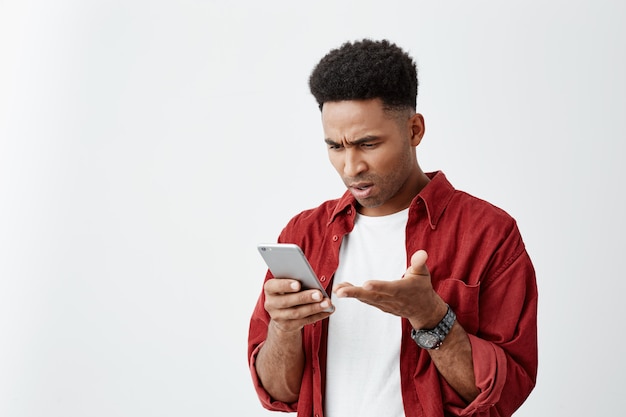 Sluit omhoog portret van de jonge knappe tan-gevilde mens met afrokapsel in toevallig wit t-shirt met rood overhemd bekijkend telefoon met verwarde uitdrukking, ruzie met vriendin.