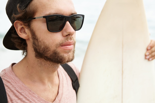 Gratis foto sluit omhoog portret van de knappe jonge mens met verwarde baard die zonnebril dragen en honkbal glb achteruit zijn witte surfplank houden en oceaan bekijken