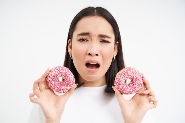 Sluit omhoog portret van droevige aziatische vrouw die op dieet is en twee geglazuurde roze donuts laat zien