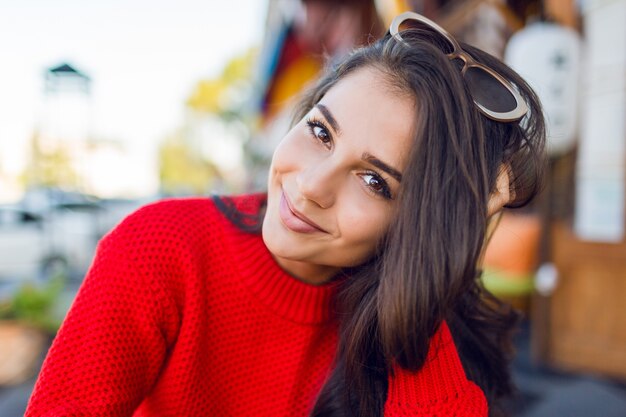 Sluit omhoog portret van elegante romantische vrouw met donkerbruine golvende haren met modieuze retro zonnebril en wed gebreide sweater. Vrouw chillen in moderne café in de ochtend en koffie drinken.