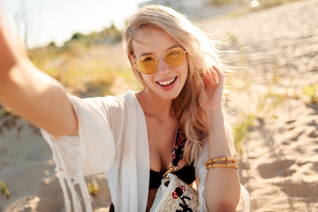 Sluit omhoog portret van glimlachende onbezorgde vrouw met blonde haren die zelfportret maken op zonnig avondstrand.