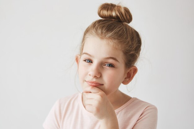 Sluit omhoog portret van grappig meisje met blond haar in broodjeskapsel, houdend vinger op kin, met geinteresseerde en foxy uitdrukking.