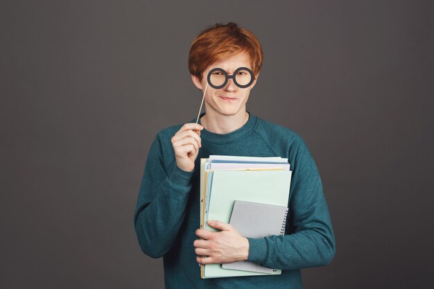 Sluit omhoog portret van grappige jonge knappe gemberkerel in groene sweater die veel notitieboekjes in hand houden, kijkend met onzekere uitdrukking door document partijglazen.