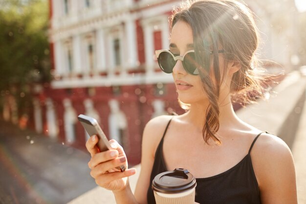 Sluit omhoog portret van jonge knappe ernstige Spaanse vrouw die met vriend babbelen dat die laat voor vergadering op celtelefoon, koffie drinken, zonnige de zomerdag buiten doorbrengen.