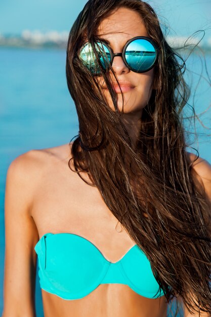 Sluit omhoog portret van modieus mooi sexy meisje in glazen en met nat haar op een zonnig strand met blauw water. Ga zonnebaden en geniet van de rust.