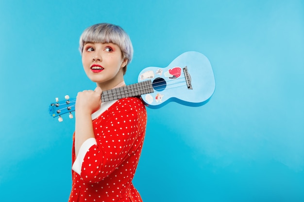 Sluit omhoog portret van mooi popachtig meisje met kort licht violet haar die de ukelele van de rode kledingsholding over blauwe muur dragen