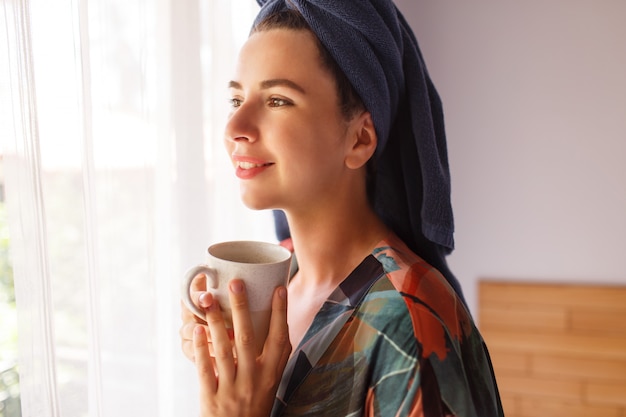 Sluit omhoog portret van mooie vrouw die in handdoek en badjasontwaken in ochtendzitting op bed wordt verpakt en thee drinkt