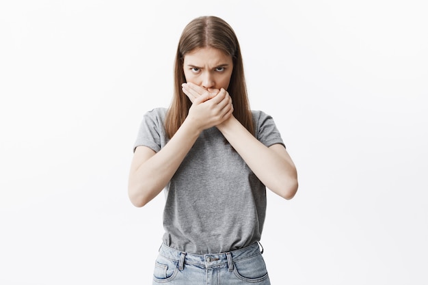 Sluit omhoog portret van ongelukkig knap eropean donkerharig meisje met lang haar in trendy kleren die mond met beide handen sluiten, met schuldige uitdrukking na het zeggen van aanstootgevend ding