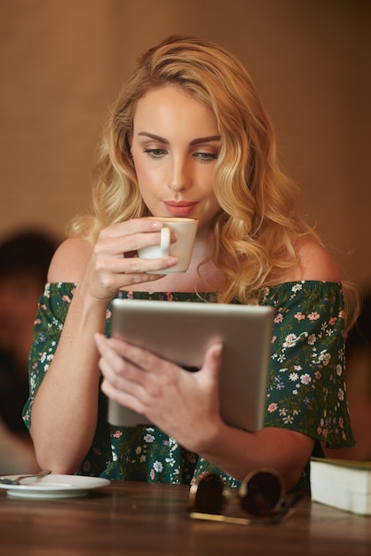 Sluit omhoog van blonde vrouw die het web op het digitale stootkussen doorbladert terwijl het drinken van koffie in een koffie