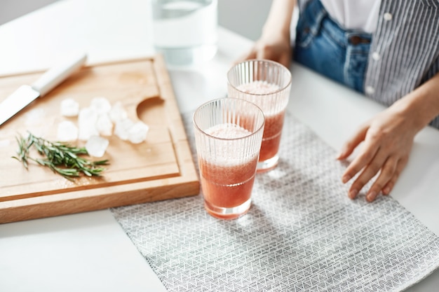 Sluit omhoog van de handenglazen van de vrouw met het dieet van de grapefruit detox smoothie rozemarijn en ijsstukken op houten bureau.