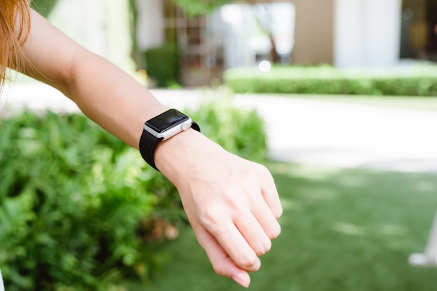 Sluit omhoog van een jonge Aziatische vrouw kijkend op haar smartwatch in de groene tuin op weekendochtend. yo