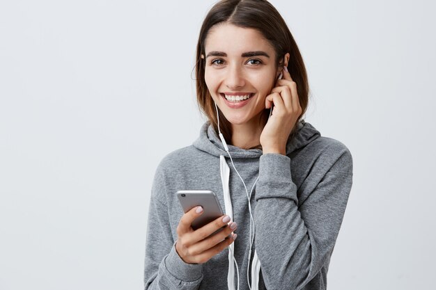 Sluit omhoog van jong vrolijk aantrekkelijk Kaukasisch meisje met donker lang haar in modieuze toevallige hoodie glimlachend met tanden, dragend oortelefoons, kijkend film op smartphone