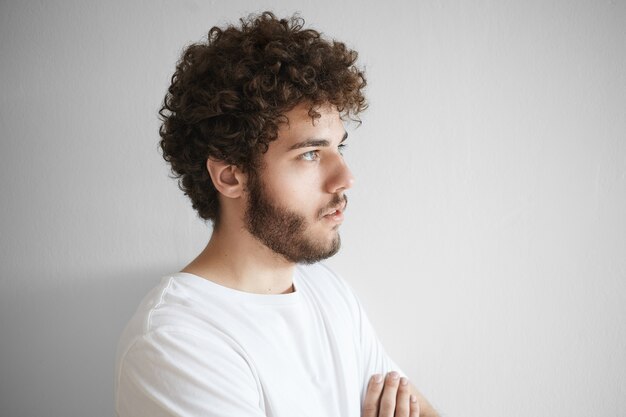 Sluit portret van aantrekkelijke jonge blanke man met krullend haar, dikke baard en mooie functies poseren geïsoleerd met lege kopie ruimte muur voor uw tekst of promotionele informatie