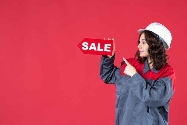 Sluit vooraanzicht van gelukkige vrouwelijke werknemer in uniform die bouwvakker draagt en verkooppictogram op geïsoleerde rode muur richt