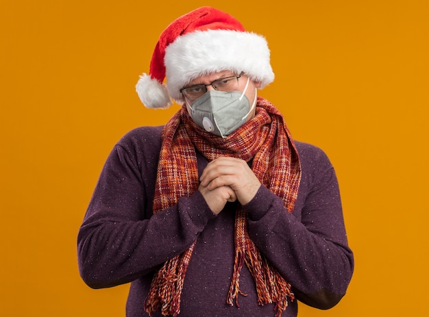 Sluwe volwassen man met beschermend masker bril en kerstmuts met sjaal om nek handen samen te houden kijken naar camera geïsoleerd op een oranje achtergrond