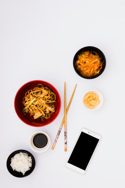 Smakelijke noedels in kom dichtbij salade; gember zuur; sojasaus en gestoomde rijst met mobiele telefoon op witte achtergrond
