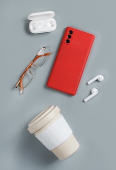 Smartphone, herbruikbare koffiekopje, draadloze oortelefoons en bril op grijze achtergrond bovenaanzicht