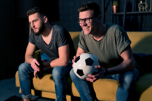 Smiley mannelijke vrienden kijken samen naar sport op tv terwijl ze voetbal vast houden