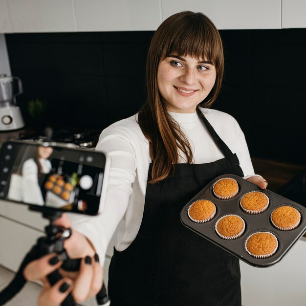 Smiley vrouwelijke blogger die zichzelf opneemt met smartphone tijdens het bereiden van muffins