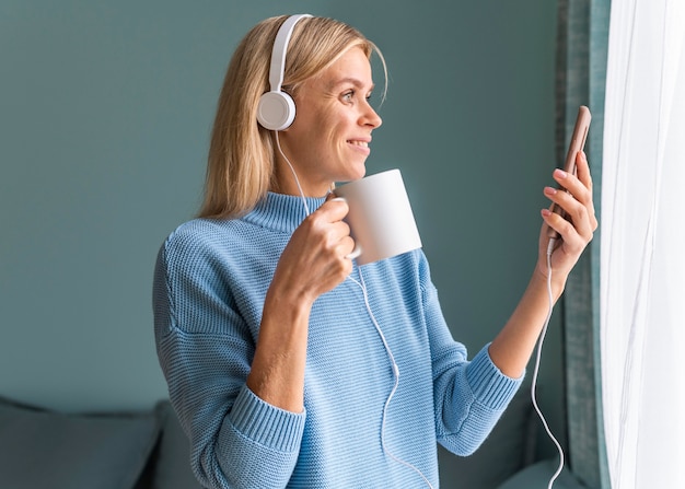 Smileyvrouw die thuis smartphone en hoofdtelefoons gebruikt terwijl het drinken van koffie tijdens de pandemie