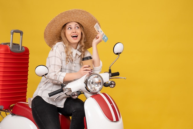 Smilingand dromerige jonge vrouw die hoed draagt en op motorfiets zit en koffie en kaartje op geel houdt