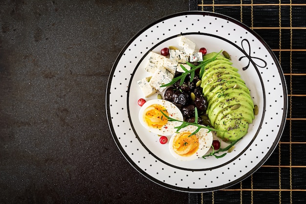 Snack of gezond ontbijt - plaat van blauwe kaas, avocado, gekookt ei, olijven op een zwarte ondergrond. Bovenaanzicht