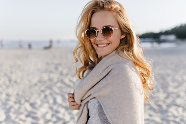 Snapportrait van krullende jonge vrouw in beige outfit en zonnebril glimlachend in het strand