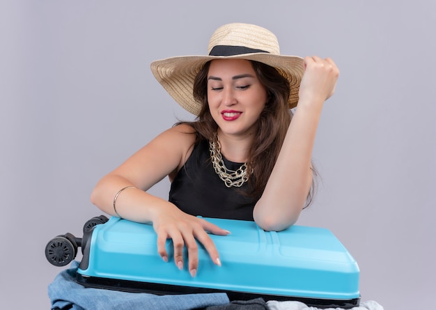 Sniling reiziger jong meisje draagt zwart onderhemd in hoed probeert koffer op witte achtergrond te sluiten