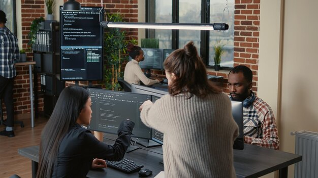 Softwareprogrammeur die potlood wijst naar broncode op computerscherm en algoritme uitlegt aan collega die naast bureau staat. Programmeurs bespreken online cloud computing met team.