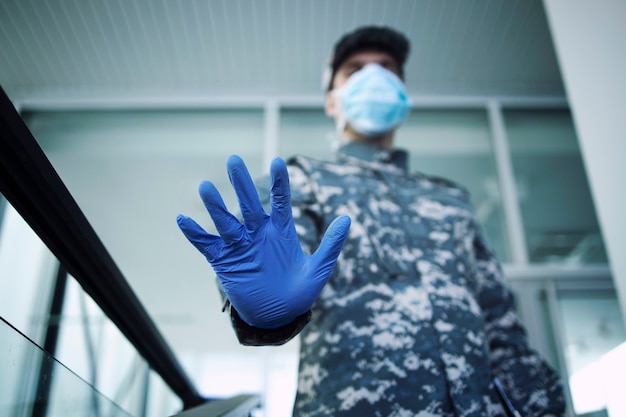 Soldaat in camouflage uniform dragen van beschermende handschoenen en masker met stopbord bij de ingang van het ziekenhuis