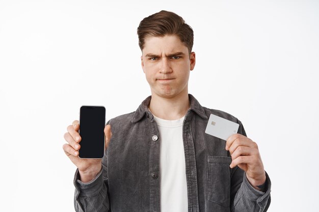 Sombere en overstuur man die creditcard met smartphonescherm toont, boos fronsend, teleurgesteld over online winkelen, financiële app, witte achtergrond. Technologie concept