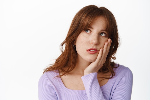 Sombere en verveelde jonge vrouw rolt met de ogen, starend naar de linkerbovenhoek met verveelde, ongeïnteresseerde gezichtsuitdrukking, moe van het wachten, staande op wit