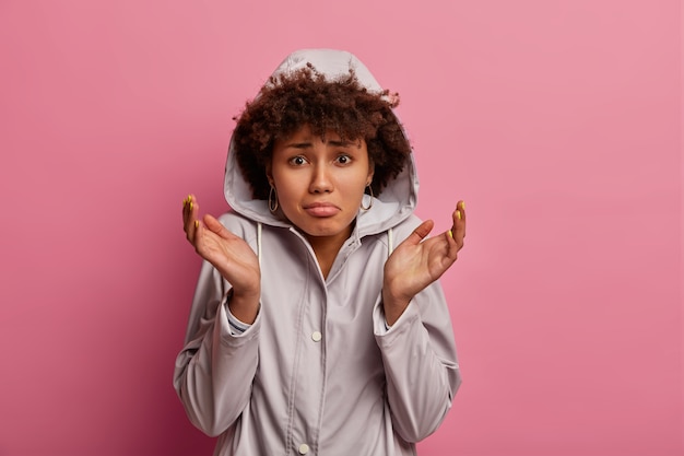 Gratis foto sombere ondervraagde vrouw wordt geconfronteerd met een gebrek aan keuzes, spreidt haar handpalmen en ziet er aarzelend uit, heeft geen idee, draagt een anorak met capuchon, voelt zich twijfelachtig en ontevreden, staat binnen. neerslachtig ellendig vrouwtje