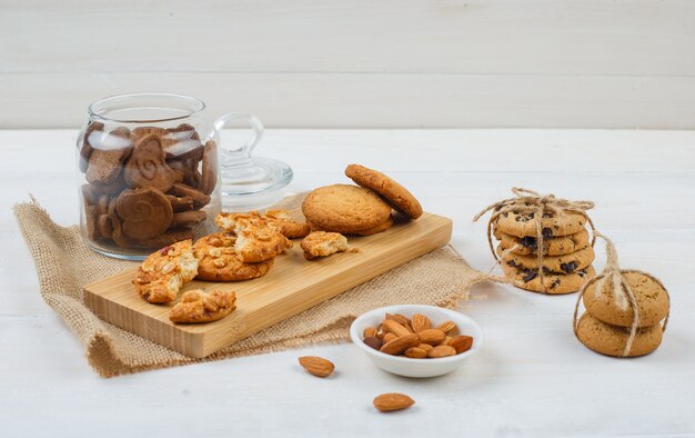 Sommige bruine koekjes met amandelen in een kom, koekjes op een snijplank en een stuk zak in een glazen pot op witte ondergrond