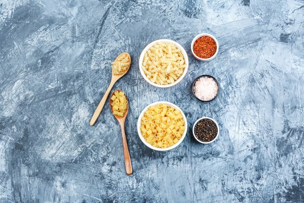 Sommige pasta met kruiden in witte kommen en houten lepels op grungy gips achtergrond, plat leggen.