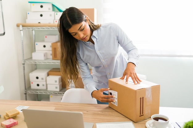 Spaanse jonge vrouw die bij haar startend bedrijf werkt en een verzendlabel op een pakket met schoonheidsproducten plakt