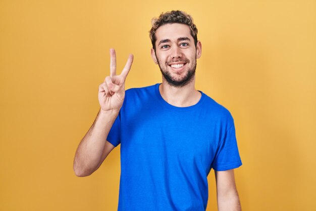 Spaanse man met baard staande op gele achtergrond glimlachend met blij gezicht knipogend naar de camera die overwinningsteken met vingers doet. nummer twee.