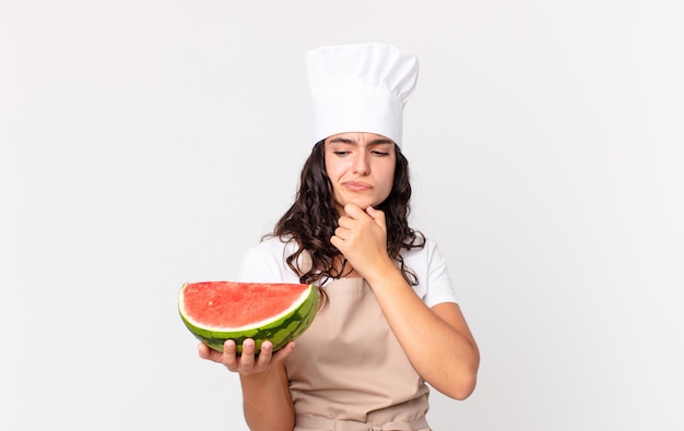 Spaanse mooie chef-kokvrouw die denkt, zich twijfelachtig en verward voelt en een watermeloen vasthoudt Premium Foto