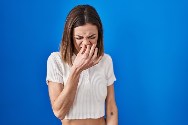 Spaanse vrouw die over een blauwe achtergrond staat en iets stinkt en walgelijk ruikt, een ondraaglijke geur inhoudt met vingers op de neus stank