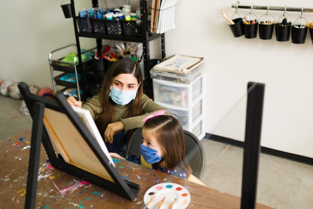 Spaanse vrouwelijke leraar instrueert een klein meisje eenvoudige schildertechnieken in een leeg canvas tijdens een kunstles. Jonge vrouw en elementair kind met gezichtsmaskers