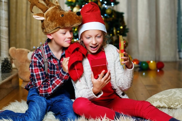 Spannende schattige jongen met kersthertenhoed en gelukkig meisje houdt een geschenkdoos in een met kerst versierde kamer.