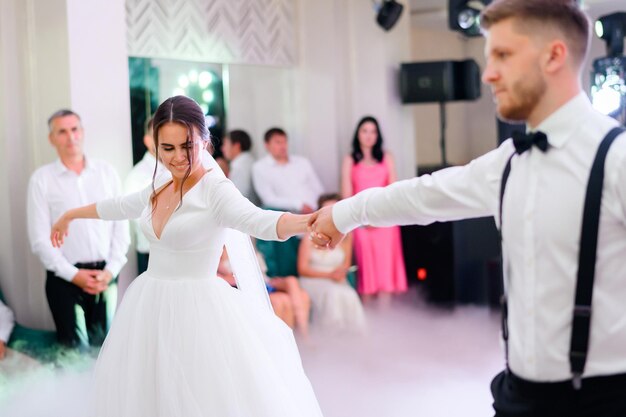 Speciaal moment van eerste dansende gelukkige bruid en bruidegom in het restaurant terwijl gasten op zoek naar mooie vrouw in elegante trouwjurk hand in hand van man huwelijksfeest