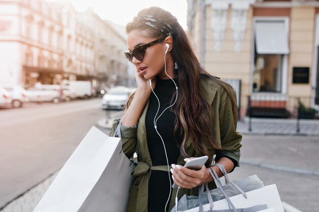 Spectaculaire Latijns-vrouw in stijlvolle zonnebril chillen in de stad en muziek luisteren in oortelefoons
