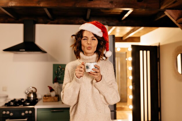 Spectaculaire mooie vrouw met krullen met een trui en een kerstmuts die een kus stuurt terwijl ze geniet van een grote kop cacao