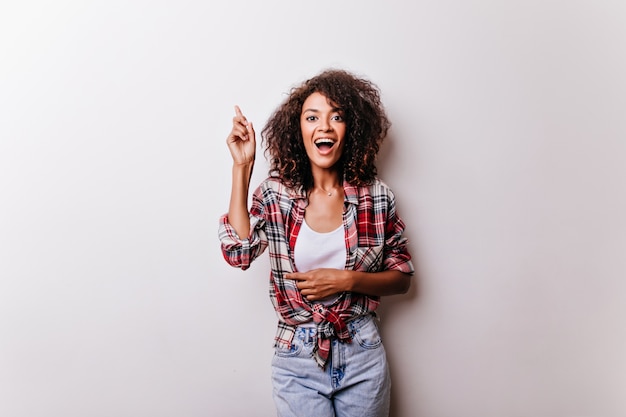 Spectaculaire vrouw in stijlvolle kleding die opwinding uitdrukt. portret van vrolijke jonge dame in geruit overhemd staande op wit.