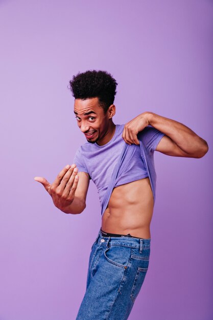 Speels Afrikaans model dat zijn t-shirt opstijgt. Portret van schattige jonge man op zoek met geïnteresseerde glimlach.