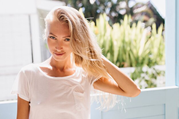 Speels blond meisje poseren in de buurt van venster. Mooie Europese dame in trendy wit t-shirt ontspannen in zonnige ochtend.