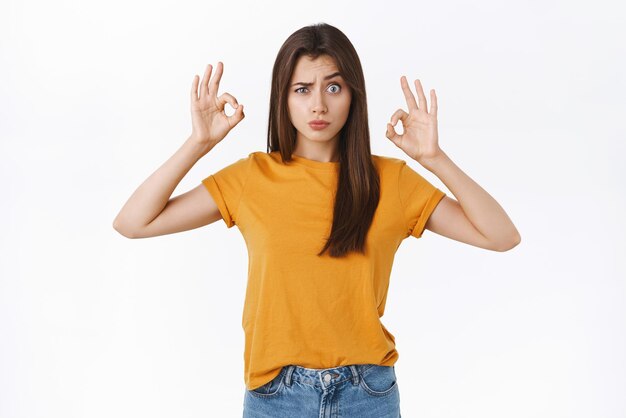 Speels dom en schattig meisje in gele t-shirt grimassen maken van grappige gezichten als het tonen van oke ok bevestiging of goedkeuring teken accepteren iets vreemds of raars zoals het witte achtergrond